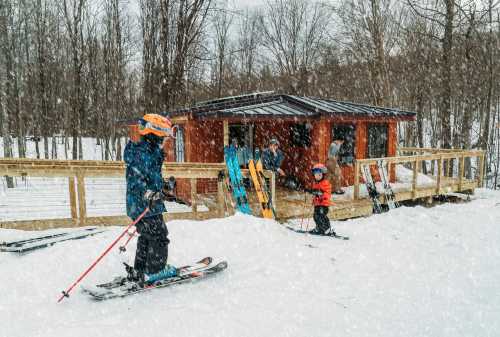 Two children skiing in a snowy landscape near a wooden cabin, with adults and ski equipment in the background.