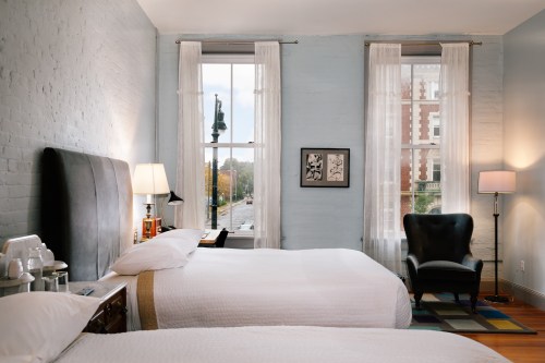 A cozy hotel room with two beds, large windows, and stylish decor, featuring a chair and a view of the street outside.