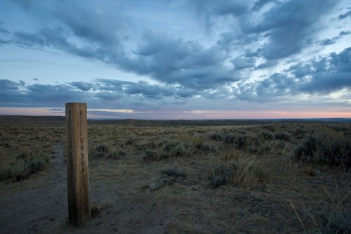A wooden post marks a trail in a vast, open landscape under a cloudy sky at dusk.