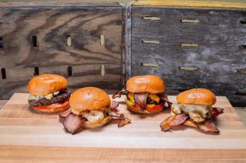 Four gourmet burgers on a wooden surface, each topped with bacon, tomatoes, and various cheeses, against a rustic backdrop.