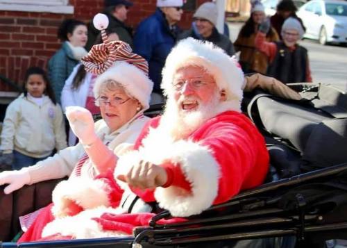 A cheerful Santa and Mrs. Claus wave from a festive carriage, surrounded by a joyful crowd in winter attire.