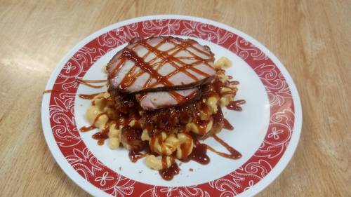 A plate of macaroni topped with pulled pork, slices of meat, and drizzled with barbecue sauce.