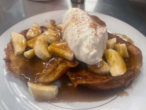 A plate of pancakes topped with sliced bananas, whipped cream, and a drizzle of syrup.