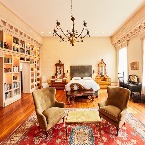 Cozy bedroom with a large bed, two armchairs, a chandelier, and bookshelves lining the walls. Warm wooden floors.