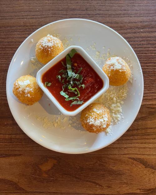 A plate with four golden fried balls and a small bowl of marinara sauce, garnished with fresh basil and grated cheese.