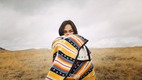 A person wrapped in a colorful blanket stands in a grassy field, with a cloudy sky in the background.