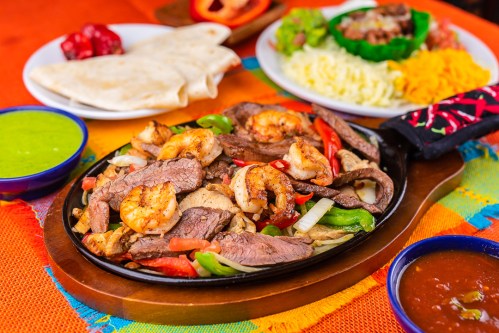 Sizzling platter of shrimp and beef fajitas with peppers, served with tortillas and sides of salsa and green sauce.