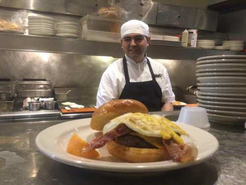 A chef stands behind a plate featuring a burger topped with bacon and a fried egg, with an orange slice on the side.