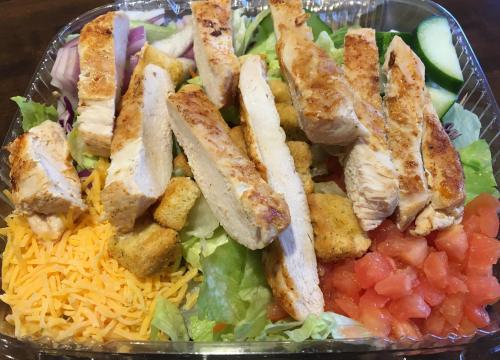 A salad with grilled chicken strips, croutons, shredded cheese, diced tomatoes, and mixed greens in a takeout container.
