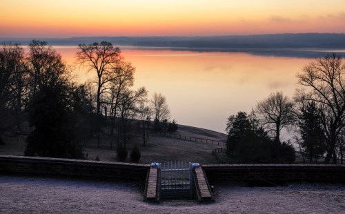 A serene sunrise over a calm lake, with trees silhouetted against the colorful sky and frost on the ground.