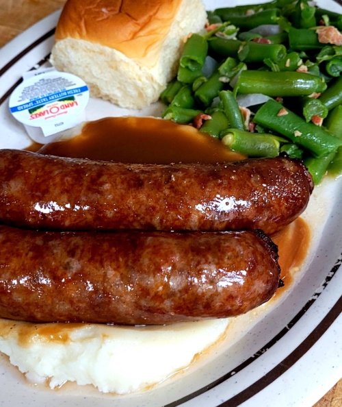 A plate with two sausages, mashed potatoes with gravy, green beans, and a roll with a butter packet.