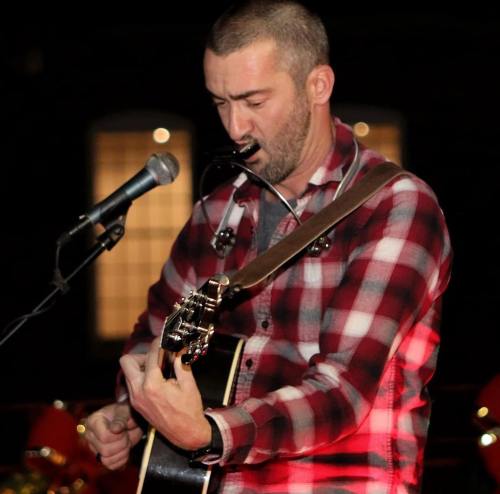 A man in a plaid shirt plays guitar and harmonica while singing into a microphone on stage.