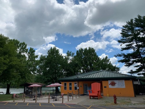 A bright orange building by a lake, surrounded by trees and picnic tables under a partly cloudy sky.