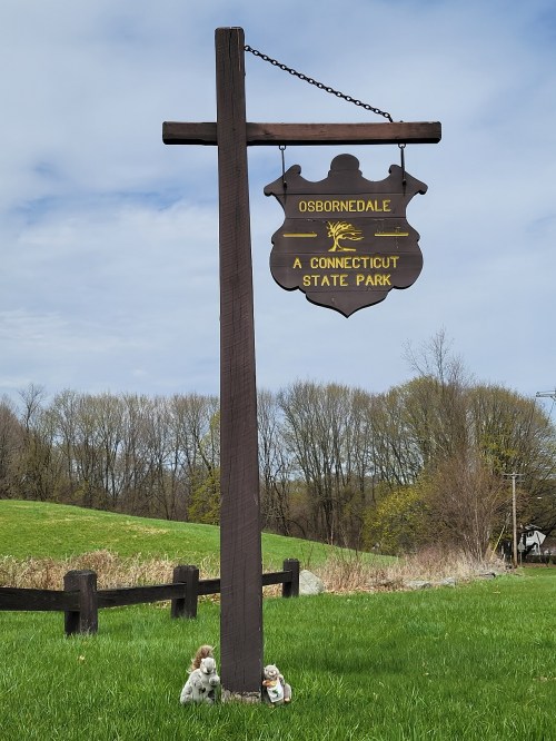 Sign for Osbornedale State Park in Connecticut, with a grassy area and trees in the background. Two small figurines at the base.