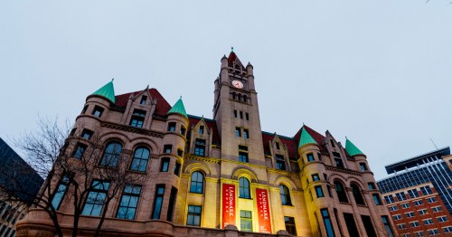 Historic building with a clock tower, featuring green-tipped roofs and banners, set against a gray sky.