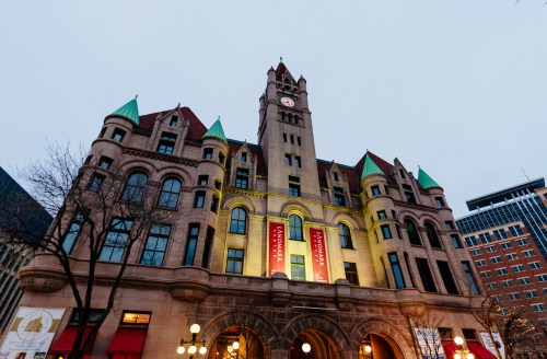 Historic building with a clock tower, featuring green roofs and illuminated banners, set against a twilight sky.