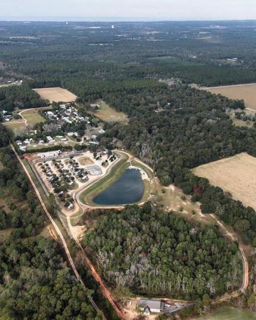 Aerial view of a rural area featuring a pond, fields, and clusters of trees with scattered buildings in the background.