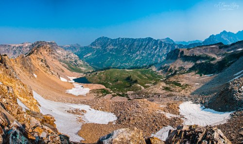 A panoramic view of rugged mountains and a green valley, with patches of snow and rocky terrain under a clear blue sky.