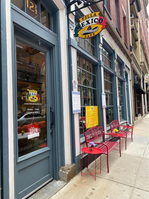 Exterior of a restaurant named "Mexico" with red chairs outside, located on a city street. Signs indicate it's open.