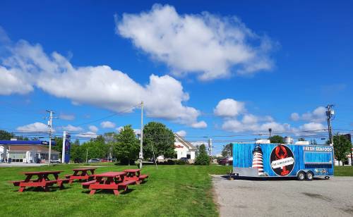 A seafood food truck parked in a grassy area with red picnic tables under a blue sky with fluffy clouds.