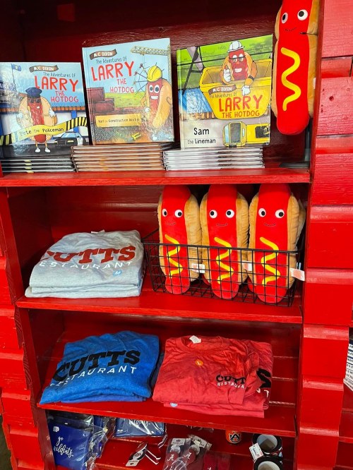 A red shelf displays "Larry the Hotdog" books, plush toys, and various restaurant-themed apparel.