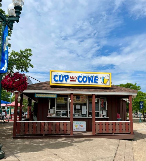 A small ice cream stand named "Cup and Cone" with a colorful sign, surrounded by trees and outdoor seating.