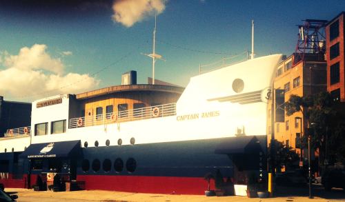 A large, white and blue ship named "Captain James" docked near buildings under a sunny sky.