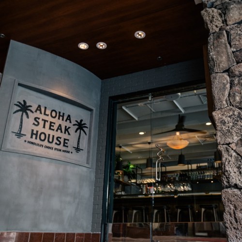 Sign for "Aloha Steak House" on a wall, featuring palm tree graphics, with a modern restaurant interior visible.