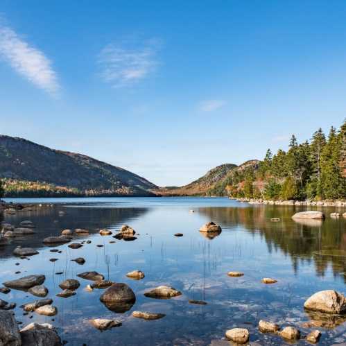 A serene lake surrounded by mountains and trees, with smooth stones scattered along the shore under a clear blue sky.