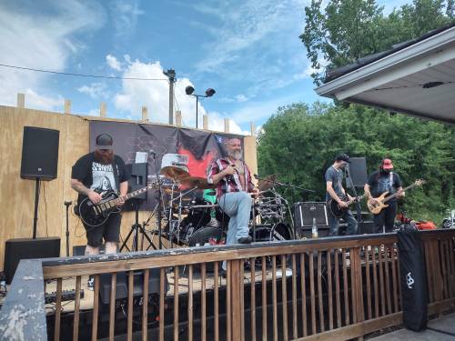 A band performs on an outdoor stage with instruments and amplifiers, surrounded by trees and a blue sky.