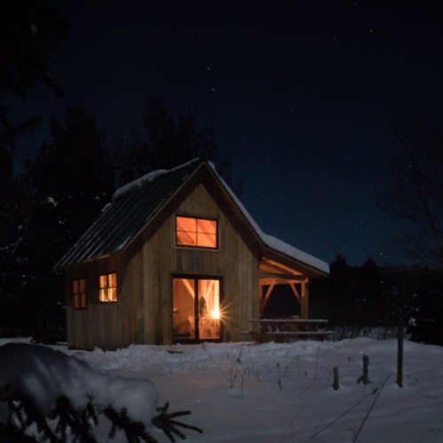 A cozy wooden cabin illuminated at night, surrounded by snow and trees under a starry sky.