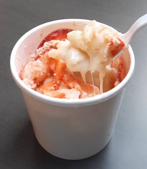 A cup of dessert featuring layers of red fruit, creamy white topping, and a sprinkle of crumbs, with a spoon.