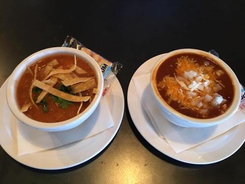 Two bowls of soup on white plates: one with tortilla strips and cilantro, the other topped with cheese and onions.