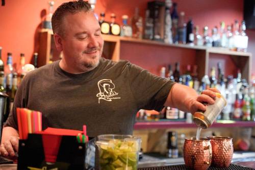 A bartender pours a drink from a can into a copper mug, smiling in a lively bar setting.