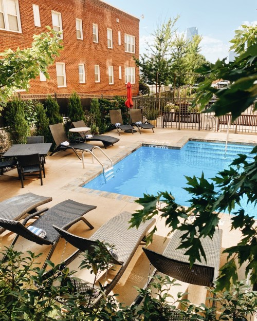 A serene pool area with lounge chairs surrounded by greenery and a brick building in the background.