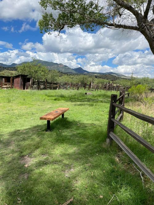 A wooden bench on green grass, surrounded by trees and a rustic fence, with mountains and clouds in the background.
