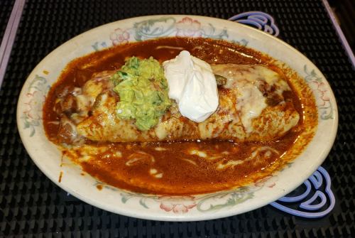 A large burrito topped with guacamole and sour cream, served in a rich red sauce on a decorative plate.