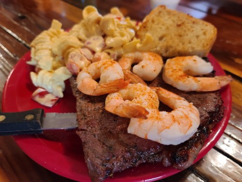 A red plate with grilled steak topped with shrimp, a side of pasta salad, and a piece of bread on a wooden table.