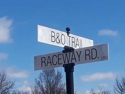 Street signs for B&O Trail and Raceway Rd against a blue sky with clouds.
