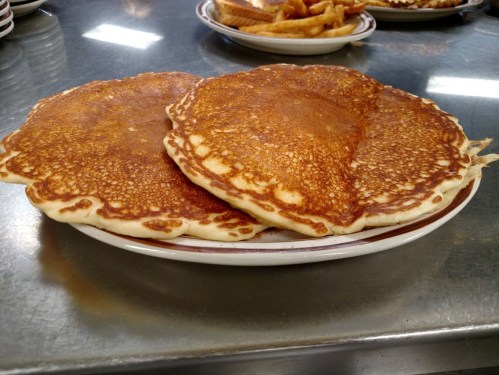 Two golden-brown pancakes stacked on a plate, with a shiny surface and a crispy edge, on a metal countertop.