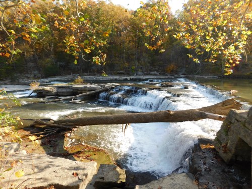 A serene waterfall cascades over rocks, surrounded by autumn foliage and a tranquil river.