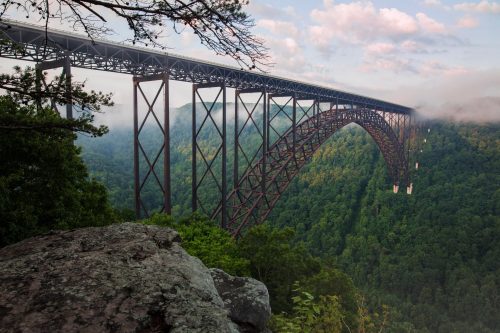A towering metal bridge spans a lush green valley, shrouded in mist under a cloudy sky.