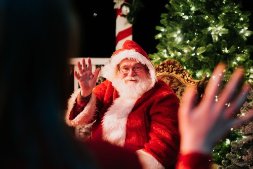 Santa Claus waves from a decorated chair, surrounded by Christmas trees and festive lights.
