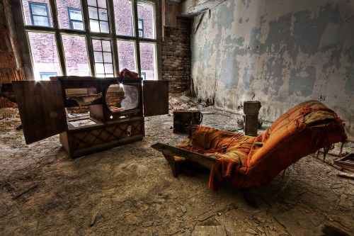 An abandoned room with a worn chair, a dusty cabinet, and peeling walls, illuminated by natural light through large windows.