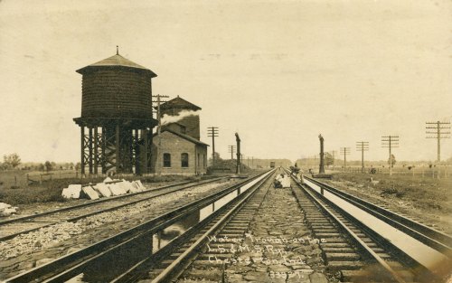 Historic railway scene featuring tracks, a water tower, and a station building under a sepia-toned sky.
