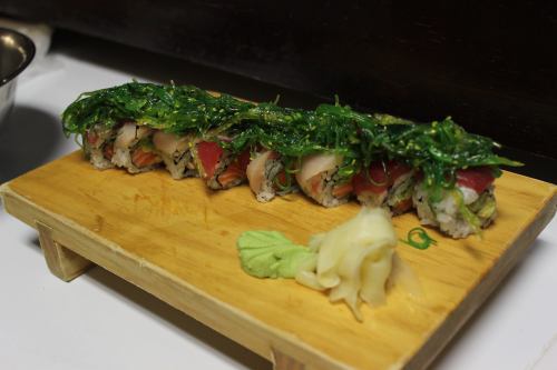 A wooden platter holds a colorful sushi roll topped with seaweed, accompanied by wasabi and pickled ginger.