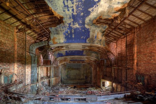 Abandoned theater interior with crumbling walls, peeling paint, and debris scattered across the floor.