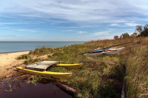 Two kayaks rest on a grassy shore by a calm lake under a blue sky with wispy clouds.