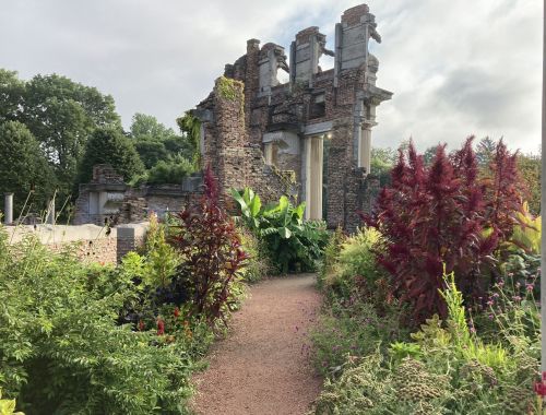 Ruins of a stone structure surrounded by lush greenery and colorful plants under a cloudy sky.