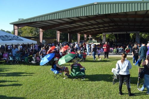 A large crowd gathers under a pavilion, with people sitting in chairs and colorful umbrellas on a grassy area.